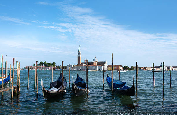 Gondolas peacefully sway on Venice's waters, facing the picturesque island of San Giorgio Maggiore, with a clear blue sky adding to the scene's tranquility.