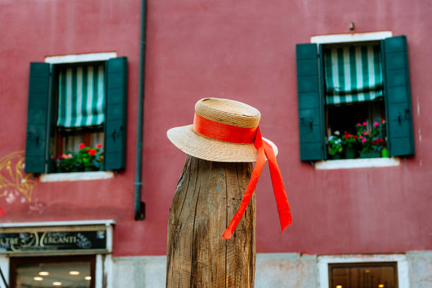 A picturesque scene of a straw hat with an orange ribbon, set on a rustic wooden post in front of a colorful Venetian building featuring green-shuttered windows and floral decor.