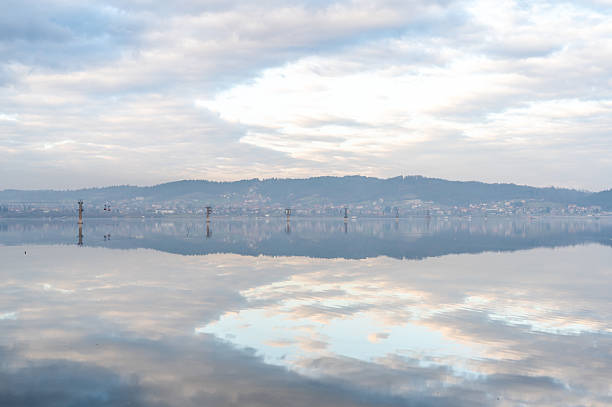Calm water surface reflecting the cloudy sky and distant hills, creating a peaceful and mirror-like effect. Cable car towers rise from the lake, adding an interesting element to the tranquil landscape. A quiet village stretches along the hillside in the background, evoking a sense of harmony between nature and human presence.