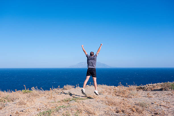 Energetic middle-aged man leaps on a hilltop overlooking the sea, symbolizing success, achievement, and personal triumph in a scenic outdoor setting.