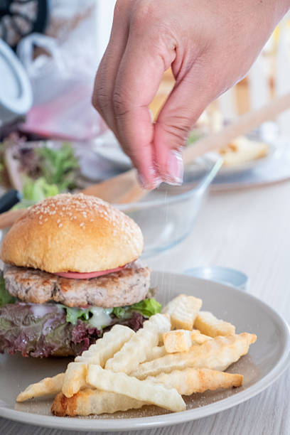 A person's hand sprinkles salt on crispy crinkle-cut french fries and a delicious homemade burger with lettuce and a patty, served on a grey plate.