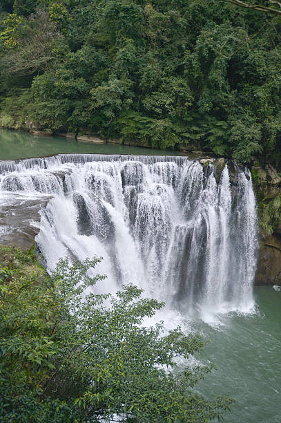 istock Powerful waterfall cascading over tiered rock formations into a vibrant green river, surrounded by dense tropical foliage and lush trees. 2230839966