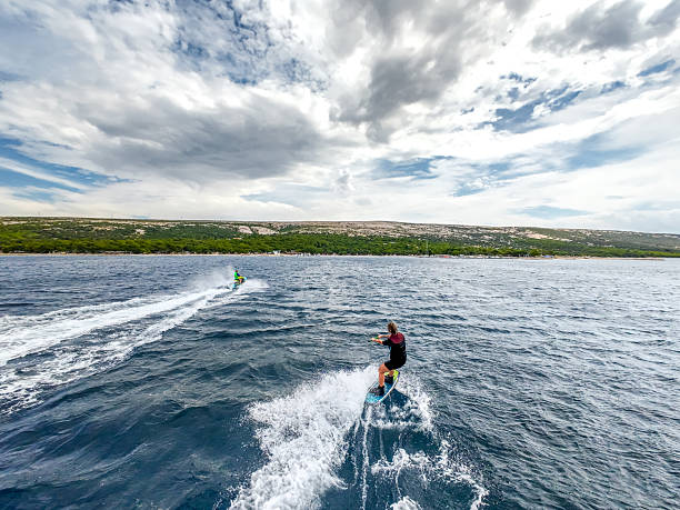 High-resolution aerial photograph capturing a female wakeboarder being towed by a jet ski on the open waters of the Adriatic Sea, along the Croatian coastline. The image showcases dynamic movement, with water spray and wake trails clearly visible from above. Dramatic cloudy skies add a striking backdrop, enhancing the sense of adventure and excitement. Taken from a drone, the perspective highlights both the skill of the wakeboarder and the natural beauty of the coastal landscape, making it perfect for illustrating water sports, outdoor recreation, travel, and active lifestyle.