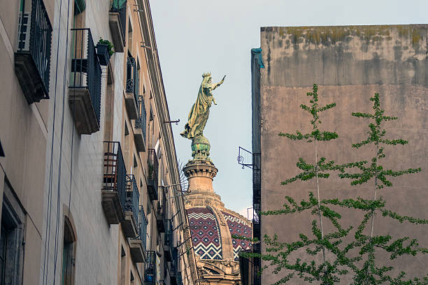 View of a historic statue rising above a colorful mosaic dome in Barcelona, Spain. The composition frames the landmark between narrow residential buildings, blending urban life with architectural heritage.