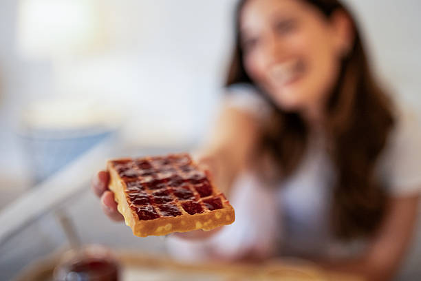 Smiling woman holding a freshly made waffle topped with jam, savoring a delightful breakfast moment filled with sweetness and joy in a cozy kitchen setting