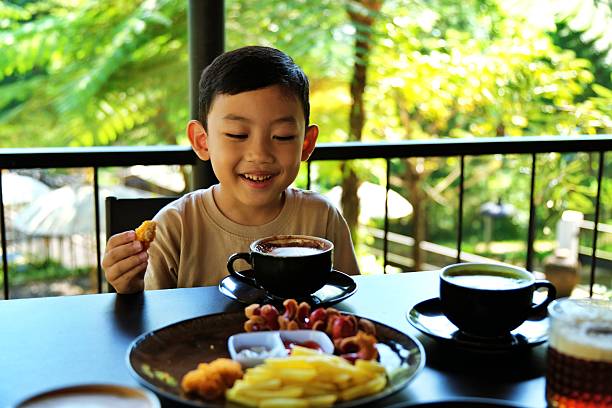 Happy young boy smiling while eating food at an outdoor cafe table with natural green view.