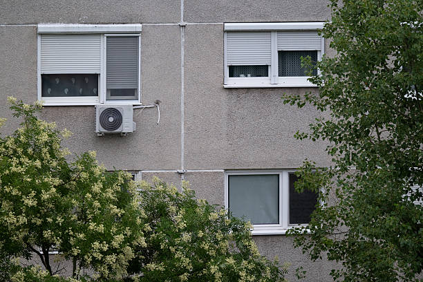 A close-up view of an apartment block facade shows multiple windows with shutters and an air conditioning unit attached to the wall. Green leafy trees partially frame the building, softening the stark concrete surface. The image reflects urban living and residential architecture.