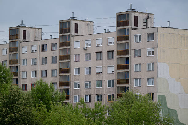 A row of aging concrete apartment buildings with multiple windows and enclosed balconies stands under a cloudy sky. The worn facade and muted colors reflect the passage of time, while green trees in the foreground add a touch of nature. The scene portrays everyday urban housing in a residential district.