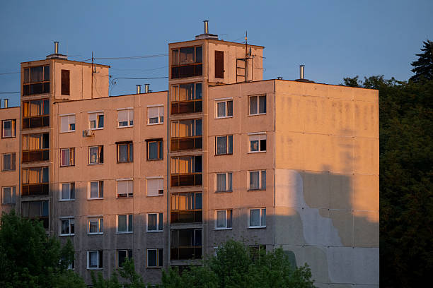 A row of old concrete apartment buildings is illuminated by the warm light of the setting sun, contrasting with the dark blue evening sky. The repetitive geometric facades and small lit windows create a sense of urban density and everyday life in a residential neighborhood. Trees in the foreground add a touch of greenery to the otherwise stark architecture.