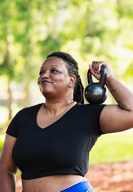 A beautiful black woman in her 30s with short hair exercising outdoors in a park on a sunny day. She is looking away, smiling as she lifts a kettlebell.