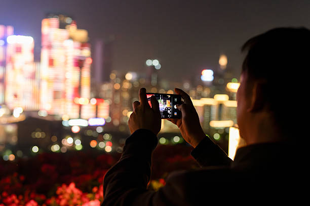 Shenzhen, China - January 17, 2019: a man holds a mobile phone with two hands to capture the nighttime light show, with illuminated buildings in the background.