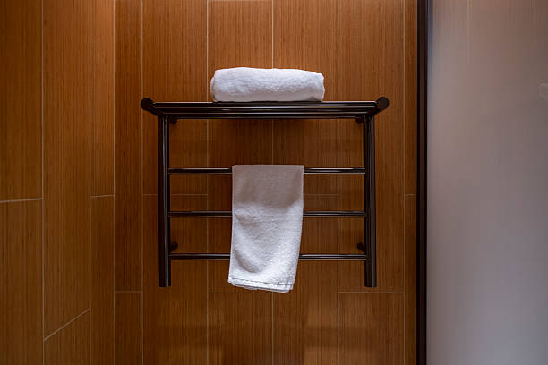 A simple yet elegant bathroom accessory setup, featuring a metal towel rack mounted on a warm-toned wooden wall. The top shelf holds a folded white towel while a hanging towel is ready for use below. The visual style conveys a sense of cleanliness and order, ideal for showcasing home decor or hospitality settings.