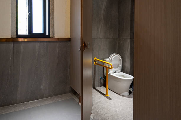 A compact bathroom featuring a toilet with a yellow grab bar, suitable for accessibility needs. The space is enclosed by wooden doors and has a window providing natural light. Visual style is utilitarian with a focus on functionality. Potential usage includes interior design inspiration or showcasing accessible bathroom features.