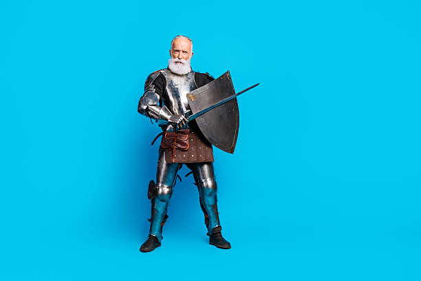 istock Senior man in medieval knight armor holding shield and sword, placed against blue background 2230915262