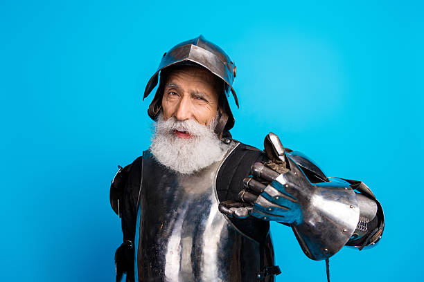 Elderly man in medieval knight armor posing on blue background exemplifying historical and mythical themes.