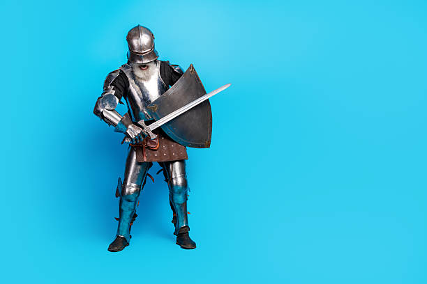 istock Mature knight in full medieval armor posing with sword and shield on a bright blue background 2230915324