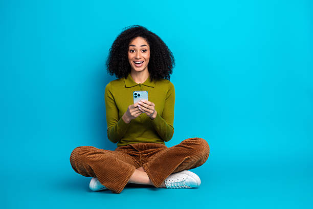 istock Cheerful young woman using smartphone while sitting cross-legged against bright blue studio background with colorful attire. 2230918117