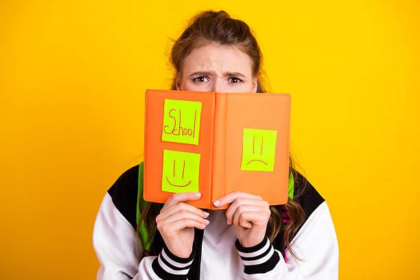 Youthful female student displays orange notebook decorated with playful sticky note faces and text, symbolizing academic life on a vivid yellow background.