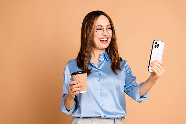 istock Confident businesswoman smiling and using smartphone while holding coffee in a blue satin blouse on beige background 2230926381