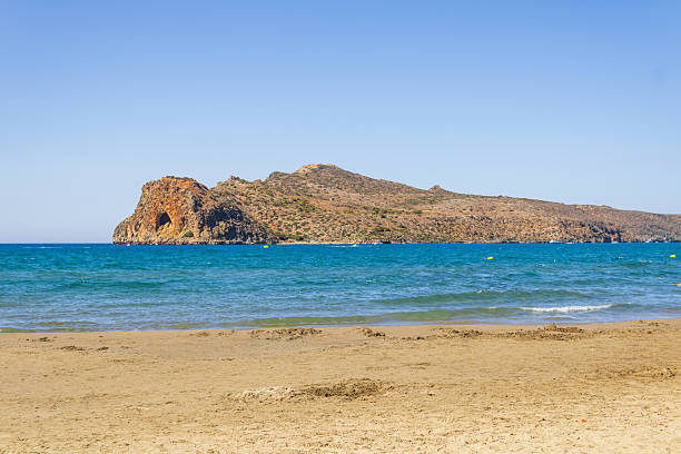 A serene beach scene with golden sand in the foreground and a rocky island rising from the turquoise sea. The calm waves gently reach the shore under a clear blue sky. The landscape evokes a sense of relaxation and natural beauty, perfect for a summer holiday destination.