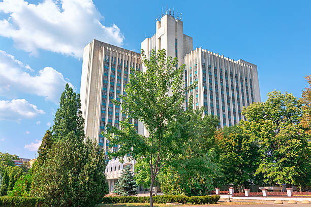 Stately government building in Chisinau stands tall behind hedges, symmetrical facade marked by vertical columns and a Moldovan flag fluttering above. Scene exudes civic formality and national pride