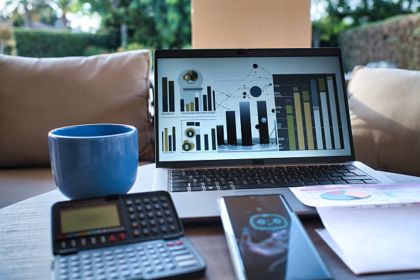 Close up of a smartphone displaying a virtual chatbot offering financial assistance. Placed beside a paper invoice, calculator, laptop with financial data, and coffee mug on a wooden terrace lit by the morning sun. Concept of digital finance, advice, and modern remote services.