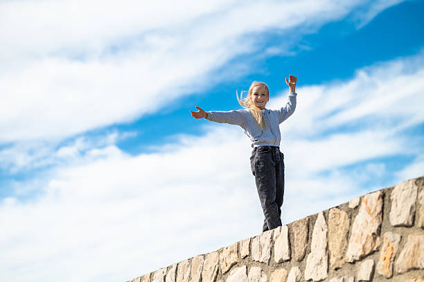 Girl balancing on stone wall arms open under clear sunny sky