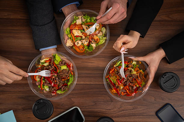 Three people are sharing healthy, colorful salads at a wooden table, suggesting a casual office lunch meeting. Top Shot