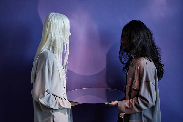 Side view studio shot of unrecognizable androgynous young woman with albinism and long-haired Indian man wearing casual clothes holding mirror looking at each other, indigo blue background