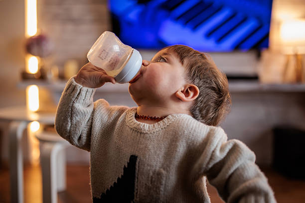 Cute baby boy wearing a warm sweater, standing near the sofa in a cozy living room, drinking milk from a bottle.
