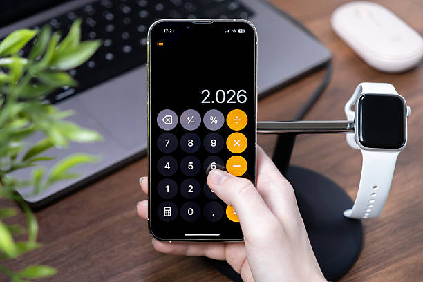 A person's hand operates a smartphone calculator app displaying 2026 for budgeting and accounting in a contemporary office setting