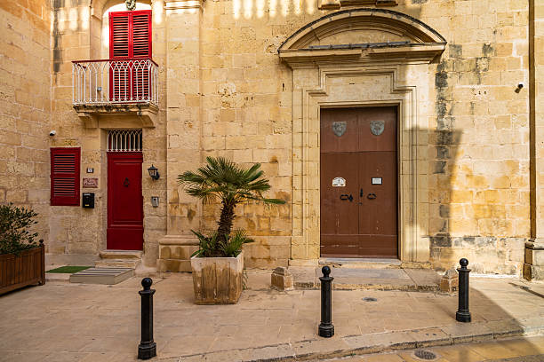 Mdina, Malta - June 22, 2021: Stone building with red doors, shutters, a small balcony, and a potted palm tree in the old town.