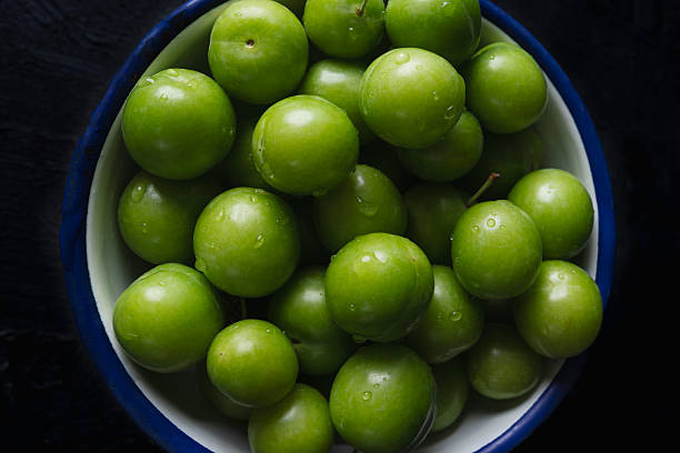 Close-up of fresh green plums in a white bowl with blue rim on a dark background. Healthy seasonal fruit rich in vitamins, perfect for diet and nutrition concepts.