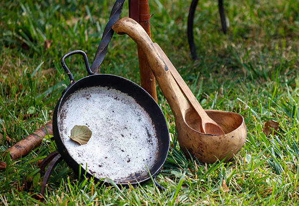 Rusty old pan and a wooden spoon are sitting on the grass. The pan is covered in dirt and has a few leaves on it. The spoon is made of wood and has a curved handle. The scene is set in a grassy area