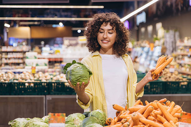 Young woman choosing between cabbage and carrots in supermarket