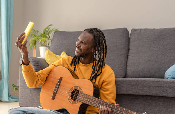 African american man with dreadlocks and yellow shirt playing guitar and using phone for streaming a live from home