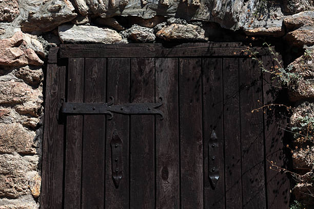 A sturdy dark wooden door blends with an aged stone wall in sunlight.
