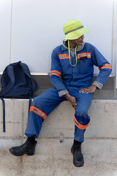 istock single african construction worker, sited outdoors , in the yard of a warehouse in workwear uniform with hat and rucksack standing in front of a warehouse wall 2230996086