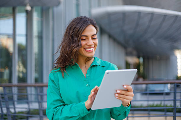istock Young businesswoman smiling while using a digital tablet, standing in front of a sleek modern office building in an urban setting 2231018383