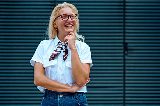 Cheerful young businesswoman with eyeglasses and a scarf, smiling and contemplating a business idea while resting her hand on her chin against a gray background