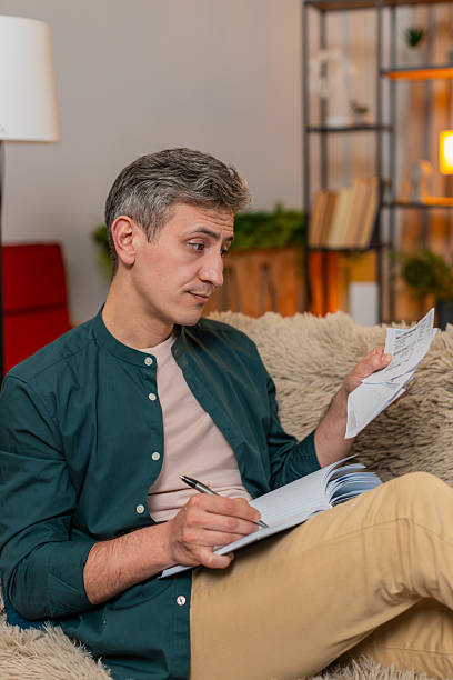 istock Man manages household budget writing numbers in notebook checking bills with serious focused look 2231023561