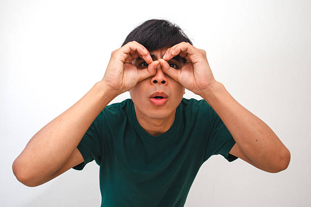 istock Portrait of a curious young Asian man looking through hand binoculars gesture with a surprised expression, isolated on a white background, perfect for creative advertising 2231031312
