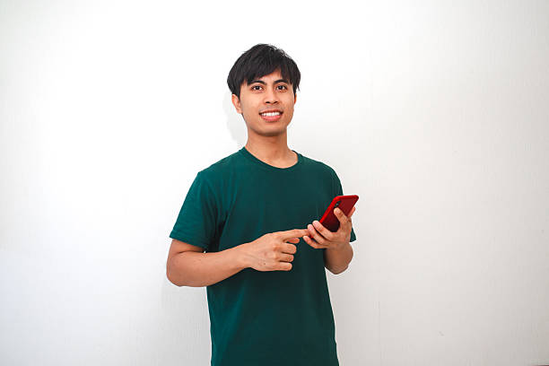 istock Smiling young Asian man holding smartphone while pressing the screen, standing on isolated white background. Perfect for advertising, tech, or lifestyle content 2231033565