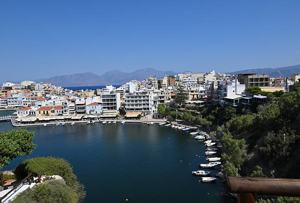 The serene lake in Agios Nikolaos, Crete, Greece, glistens under a cloudless blue sky, surrounded by charming white and pastel-colored buildings. Framed by lush green trees in the foreground, the calm water reflects the vibrant coastal scenery, creating a picturesque Mediterranean view that blends natural beauty with Greek island charm