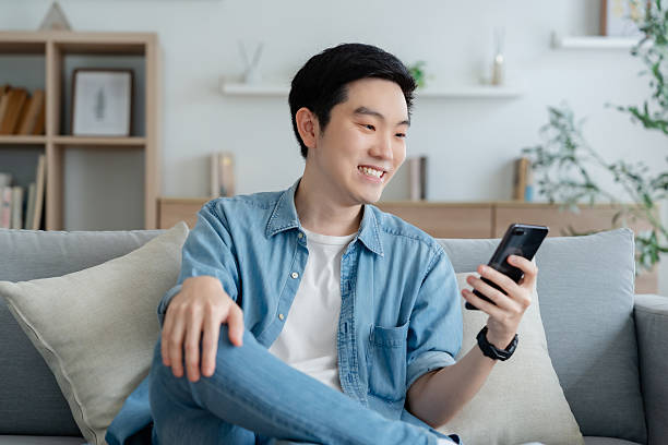 istock Smiling handsome young Asian man sitting on cozy couch, using funny mobile apps in living room. man at home, doing online shopping, messaging friends, posting on social media 2231052805