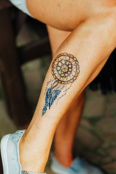 A vibrant dreamcatcher tattoo with orange and blue feathers adorns a woman's calf, showcasing intricate detail and cultural symbolism