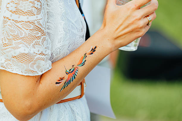 A woman's arm displays a vibrant, temporary hummingbird tattoo with vivid orange, blue, and green feathers, set against a soft outdoor backdrop