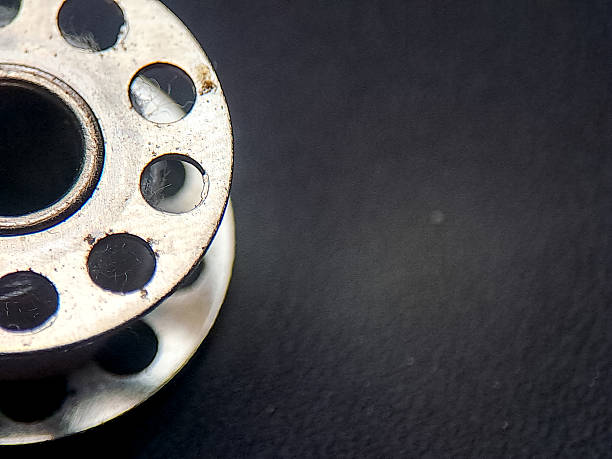A silver sewing bobbin casting geometric shadows on a dark surface, showing a stark contrast between metal and a flat plane, with a minimalist, industrial aesthetic displayed.