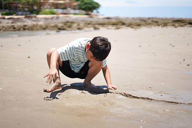 Boy playing in the sandbox with a yellow toy truck and two buckets, surrounded by dry sand and grass on a sunny day, captured during early childhood play activity.