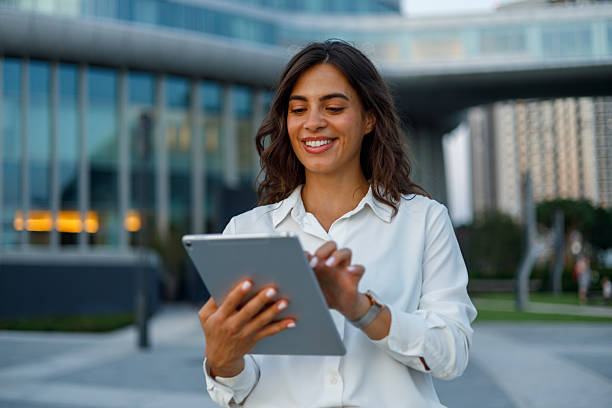 istock Young businesswoman smiling while using digital tablet outside modern office buildings in urban setting 2231068937